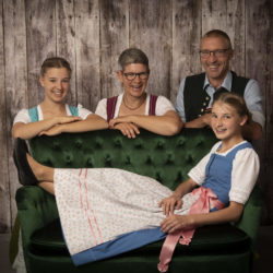 Familienfotografie: Familie in Tracht (3 Personen) schaut hinter einem grünem Sofa hervor, ein Mädchen liegt auf dem Sofa.